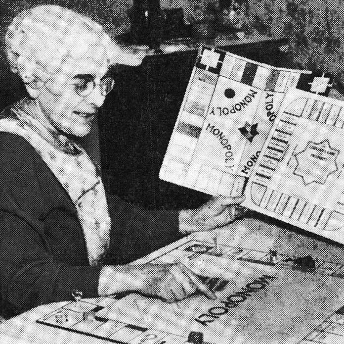 A pictures of Elizabeth Magie holding up a Monolpoly board next to her Landloard's board
